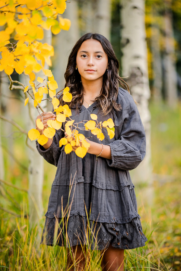 a young girl in a gray dress holding leaves in an aspen grove during the fall