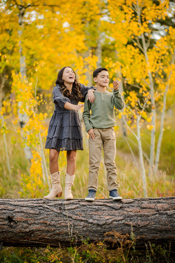 a young girl and boy standing on a fallen tree in an aspen grove during the fall
