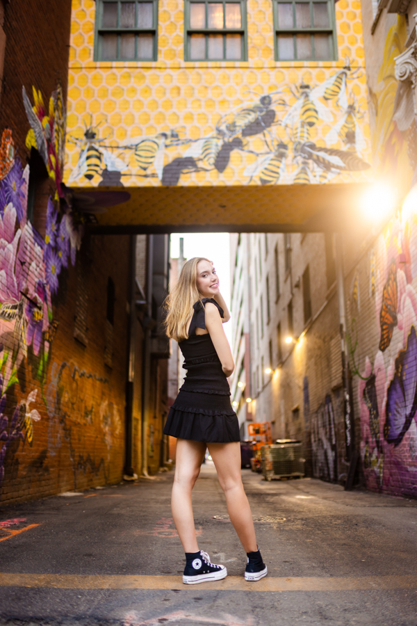 A girl in a black dress looking back at the camera while stanind in the middle of an alley