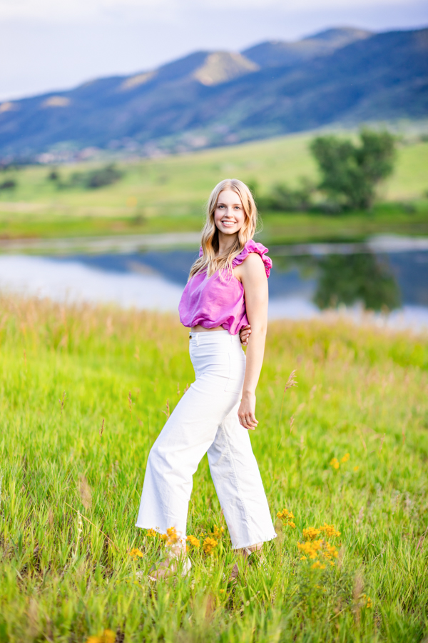 A girl in a pink top and white pants posing in front of a pond in the mountains