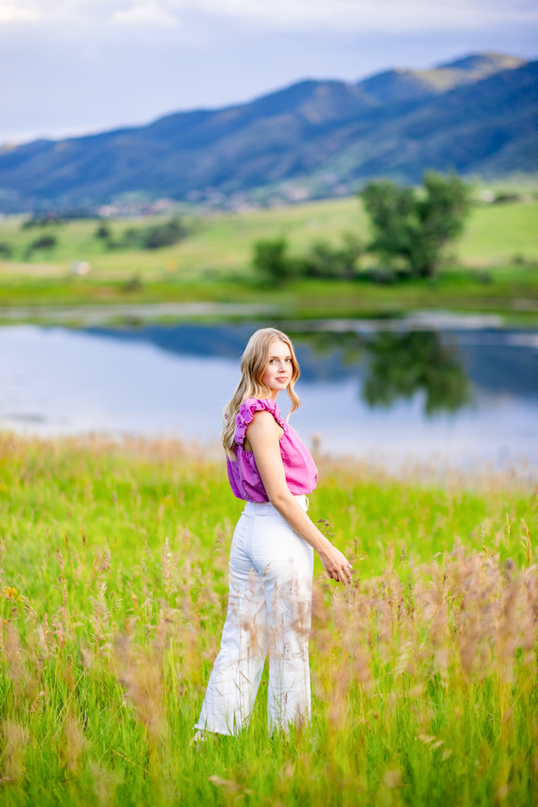 A girl in a pink top and white pants looking back in front of a pond in the mountains